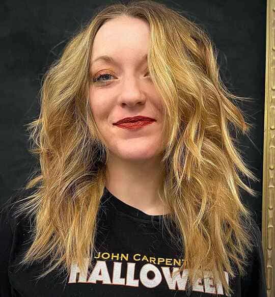 A woman with long blonde layered hair with curtain bangs and  wearing a halloween t - shirt 